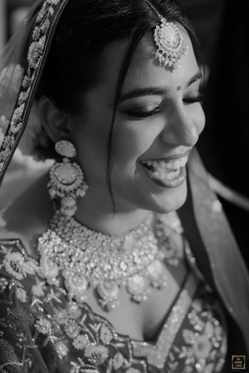 Black and white close-up of a smiling Indian bride wearing traditional jewelry, including a maang tikka on her forehead, earrings, and a heavily embroidered saree or lehenga.