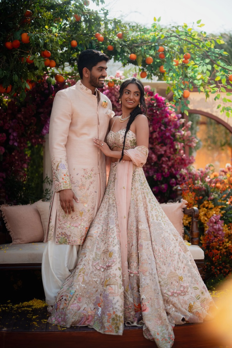 A young couple dressed in traditional Indian wedding attire, standing in a garden with orange fruit trees, pink and purple flowers, and greenery.