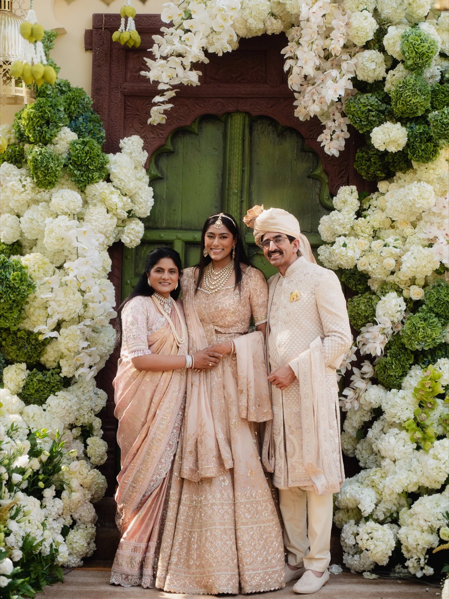 A group of four people dressed in traditional Indian wedding attire, standing in front of a floral archway with white flowers and greenery, on a green wooden door background.