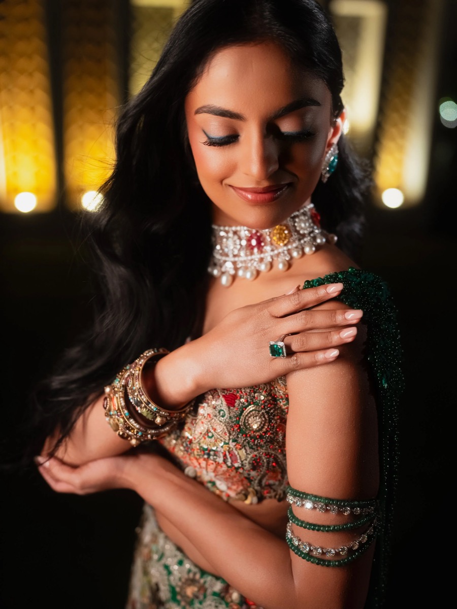 A woman with long black hair in traditional colorful attire, adorned with jewelry, closed eyes, and smiling, standing in a dimly lit setting with warm lights in the background.
