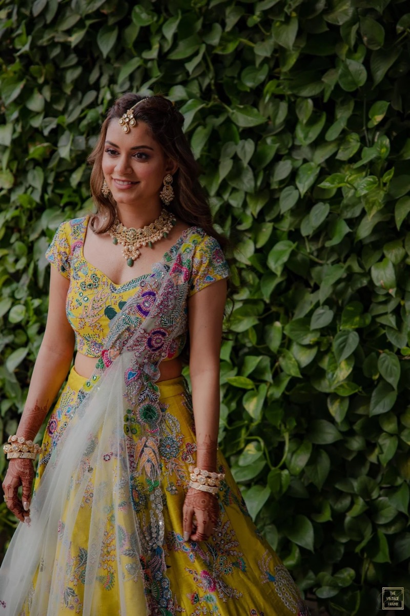A woman dressed in a colorful embroidered traditional Indian outfit standing in front of a lush green leafy background.