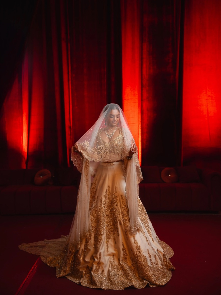 A woman in a gold wedding dress with intricate embellishments, wearing a veil,standing in front of a red curtain with dim lighting.