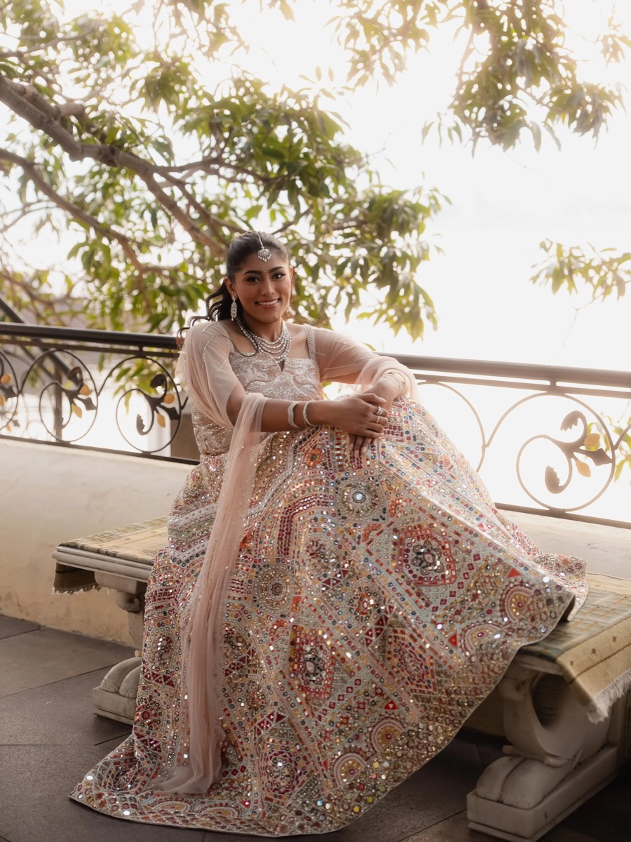 A woman dressed in traditional, intricately embroidered Indian attire sitting on a bench on a balcony with a metal railing and leafy trees in the background.