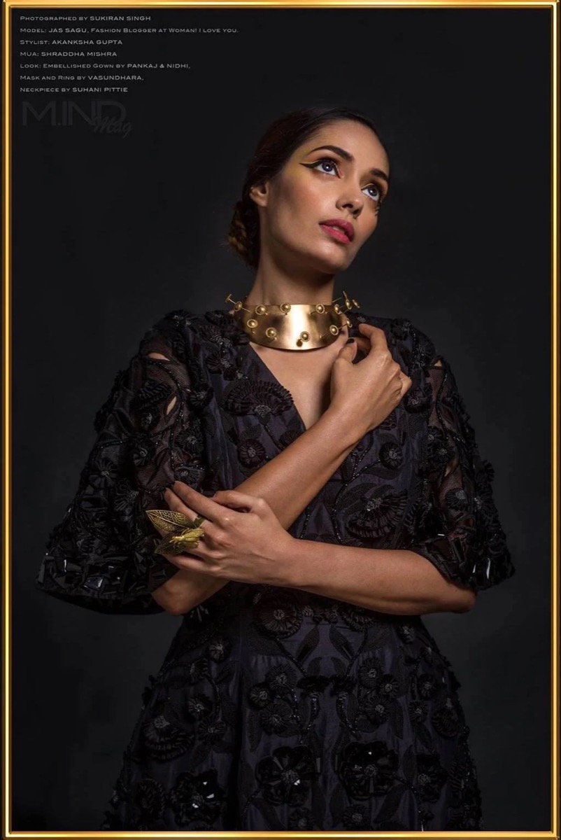 A woman wearing a black embroidered dress, a gold choker necklace with dangling circular elements, and gold statement rings. She has makeup with winged eyeliner and subtle lipstick. She poses with one hand near her chest, the other arm across her bod