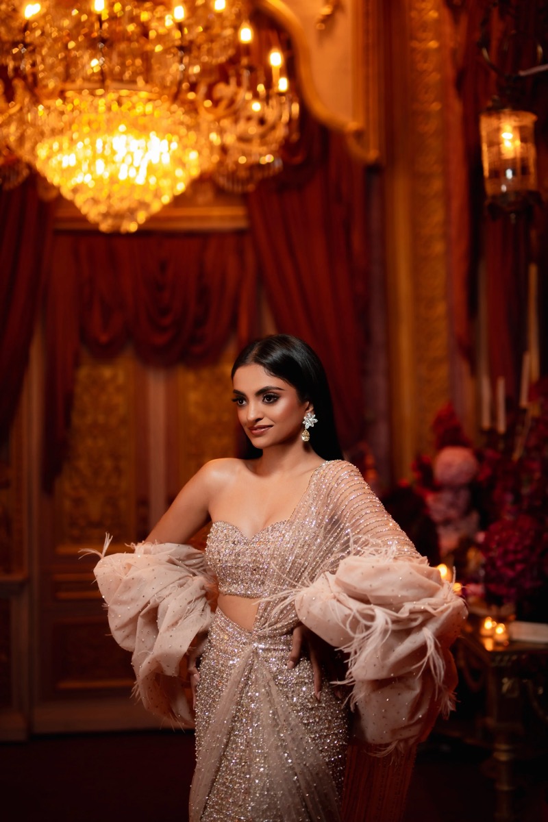 A woman in an elegant, sparkling gown stands in a luxurious, ornately decorated room with chandeliers and floral arrangements.